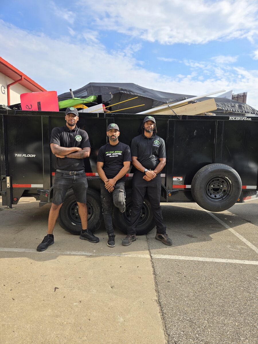 Three NYA Junk Removal crew members standing in front of their high-wall dump trailer, loaded to the rail with a finished job.